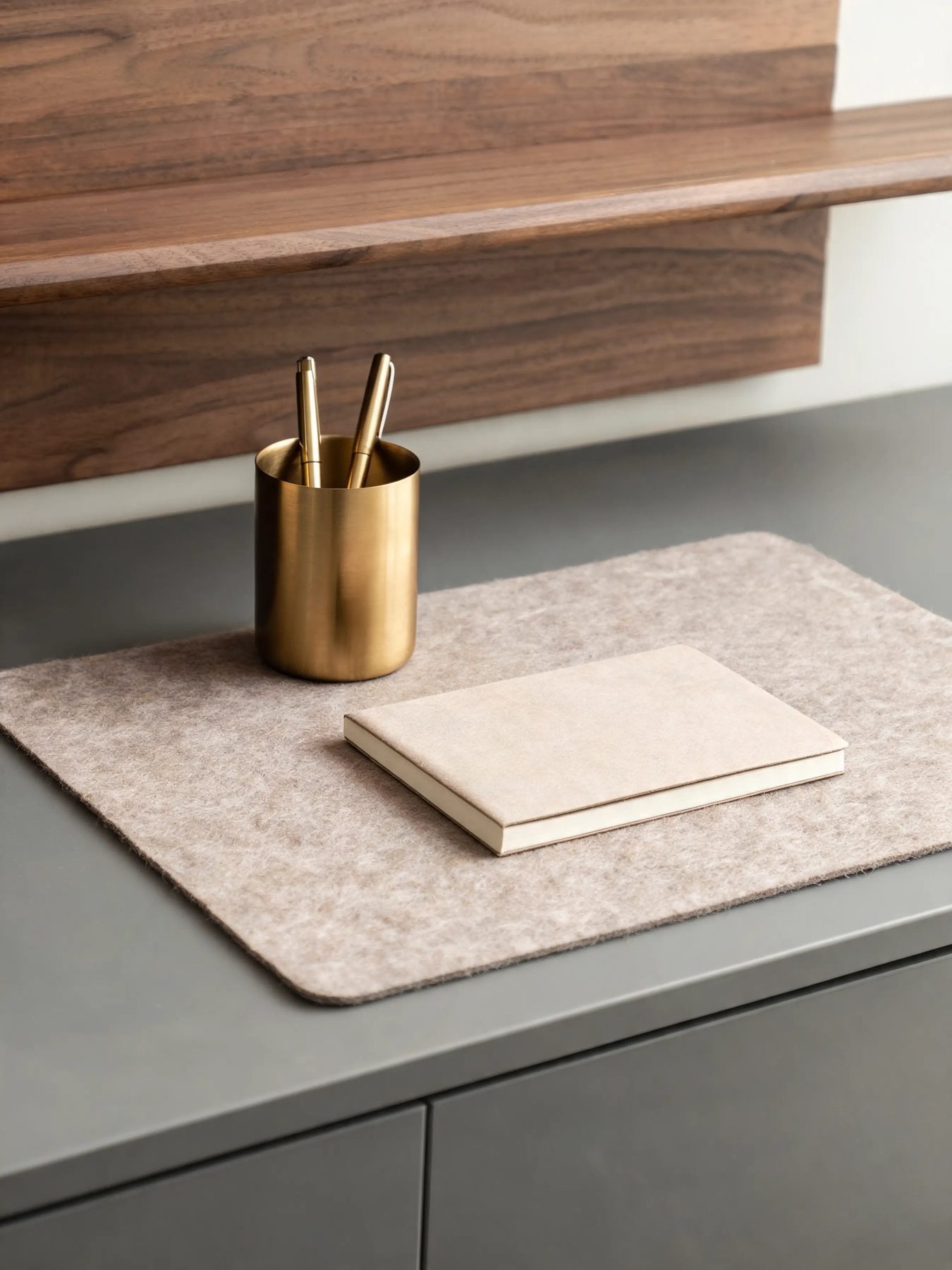Walnut desk shelf with a felt mat and a brass pen cup on a matte grey desk